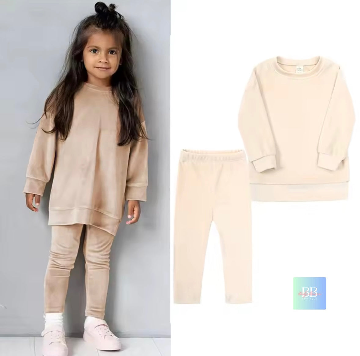 Child wearing a beige outfit with matching pants, standing against a gray wall.