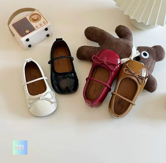 Four children's shoes in white, black, red, and brown with a small teddy bear toy on a light background.