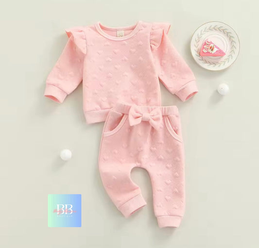 Pink baby outfit with ruffled sleeves and bow details on a light gray background.