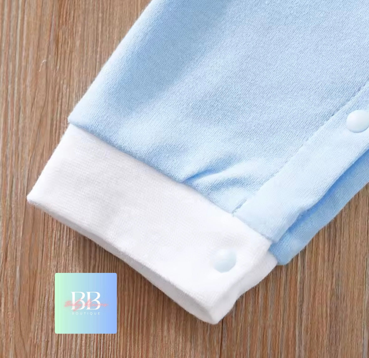 Folded baby onesie with blue top and white bottom on a wooden surface, featuring a brand logo in the corner.