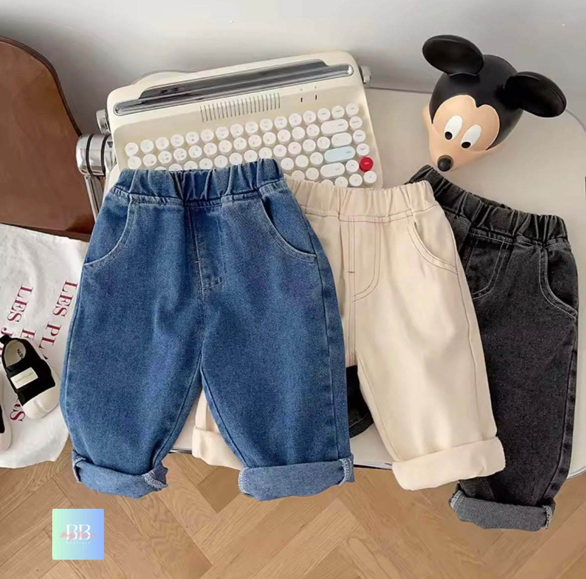 Three pairs of children's pants in blue, beige, and black on a desk with a typewriter and toy.