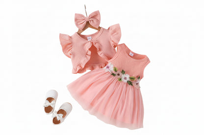 Pink baby dress with floral details, matching headband, and white shoes on a textured surface.