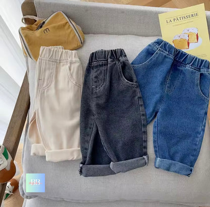 Three pairs of children's pants in beige, gray, and blue on a gray cushion with a yellow book titled 'La Pâtisserie'.