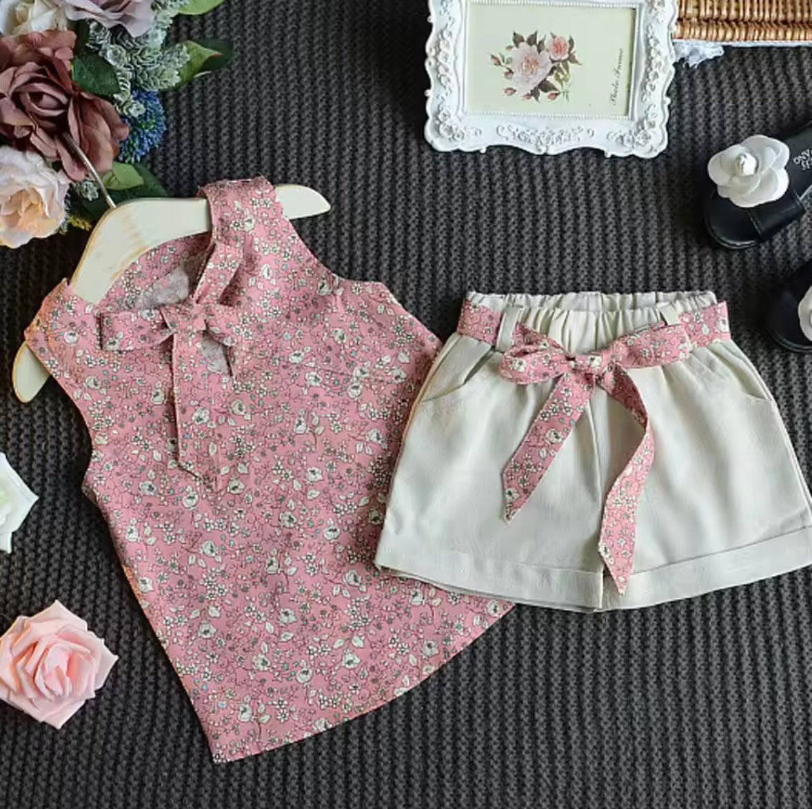 Pink floral top and white shorts with a pink ribbon on a textured surface with flowers and a picture frame.