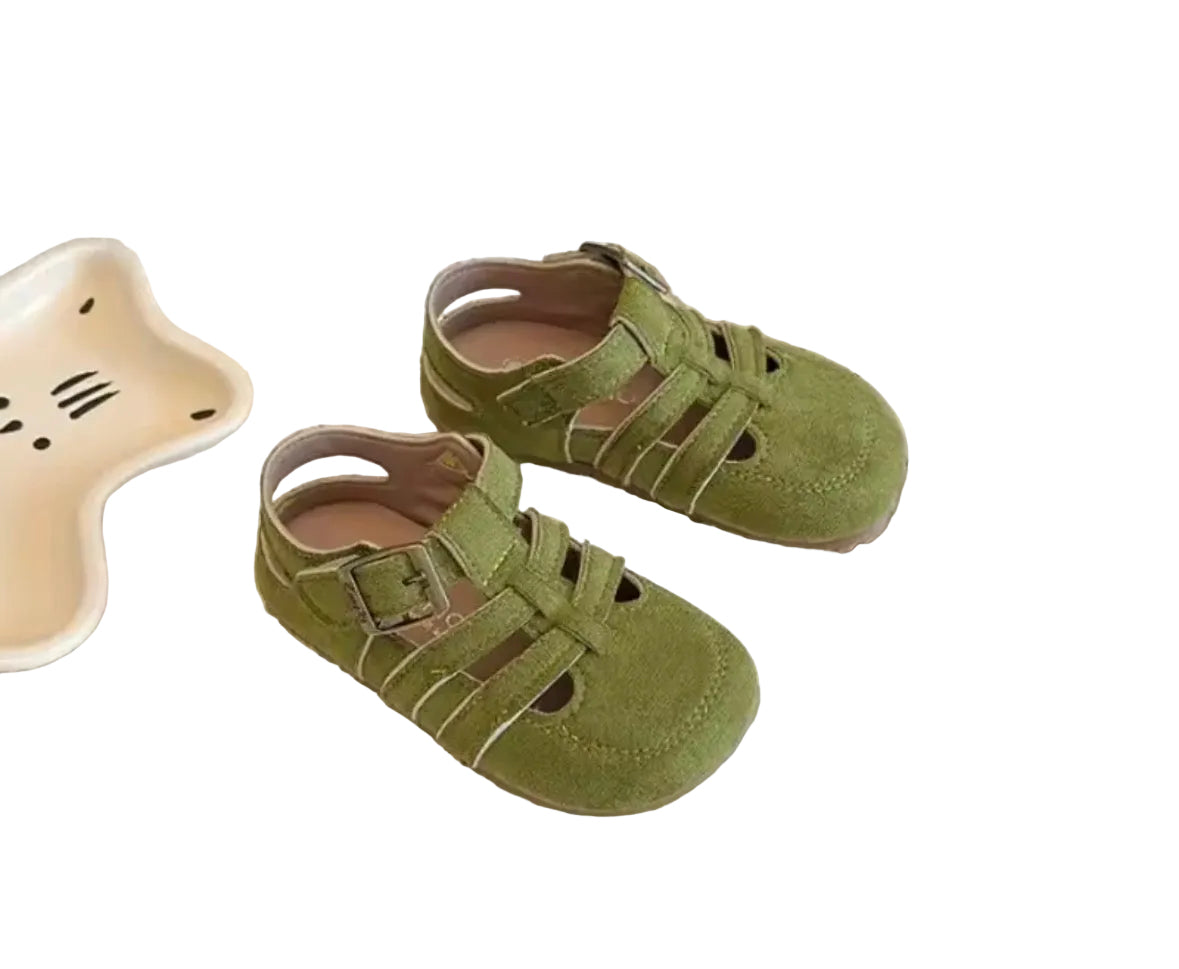 Green children's sandals on a light surface with a bottle and dish in the background