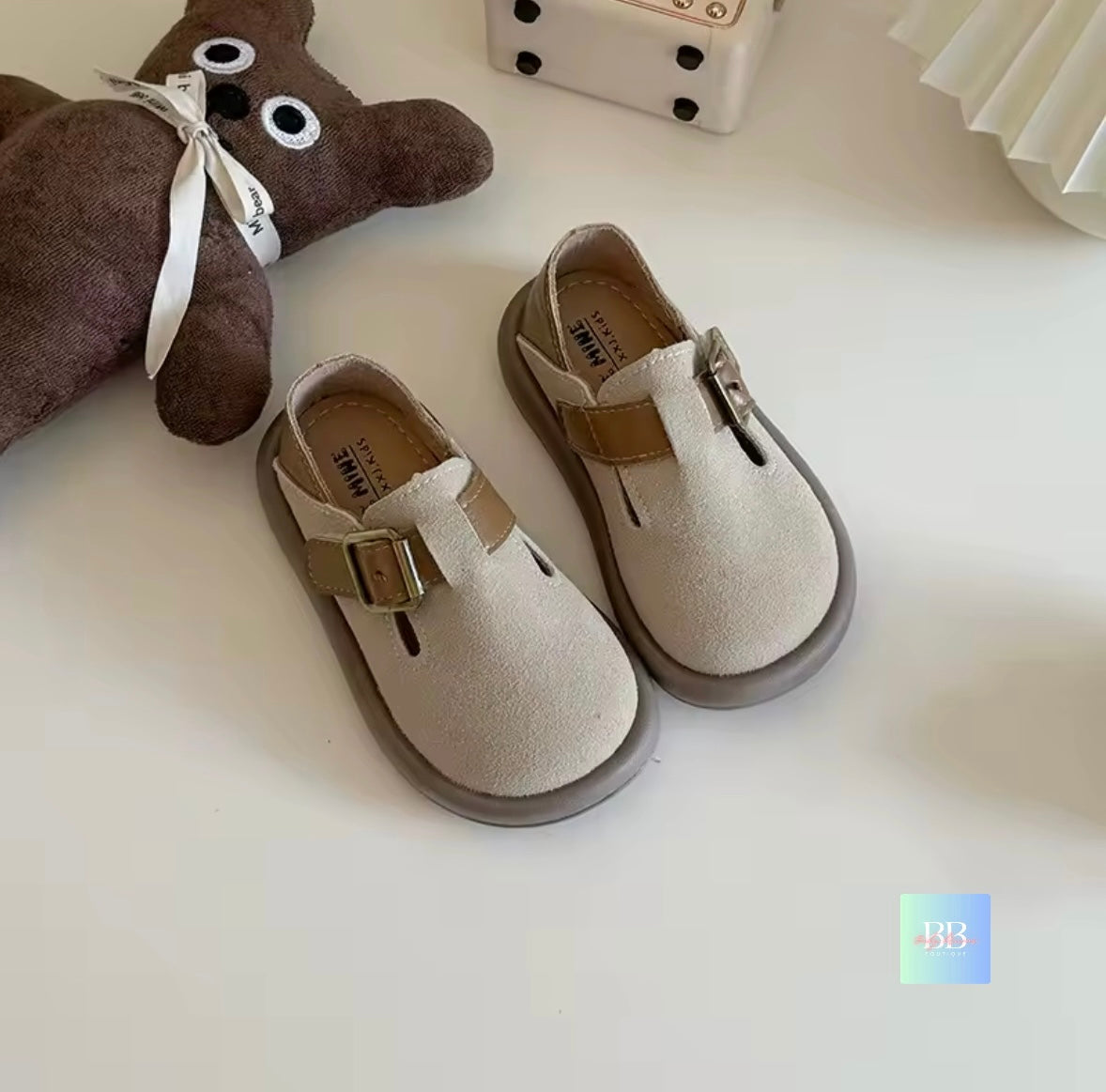 Children's beige shoes with brown buckles on a white surface, next to a brown teddy bear.