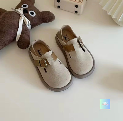 Children's beige shoes with brown buckles on a white surface, next to a brown teddy bear.