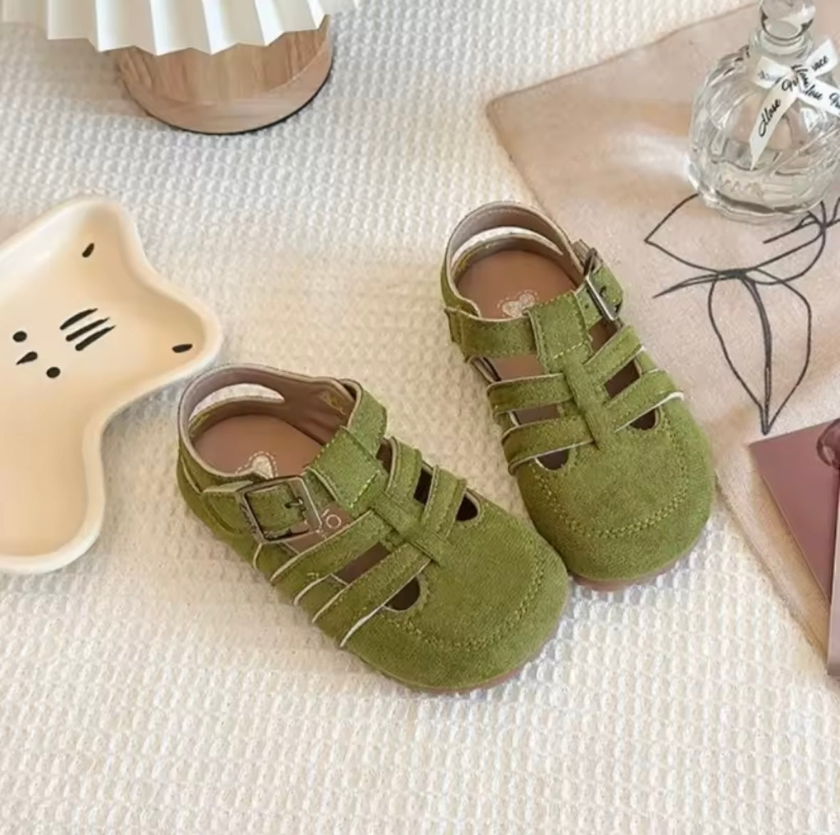 Green children's sandals on a textured surface with a star-shaped object and bottle in the background.