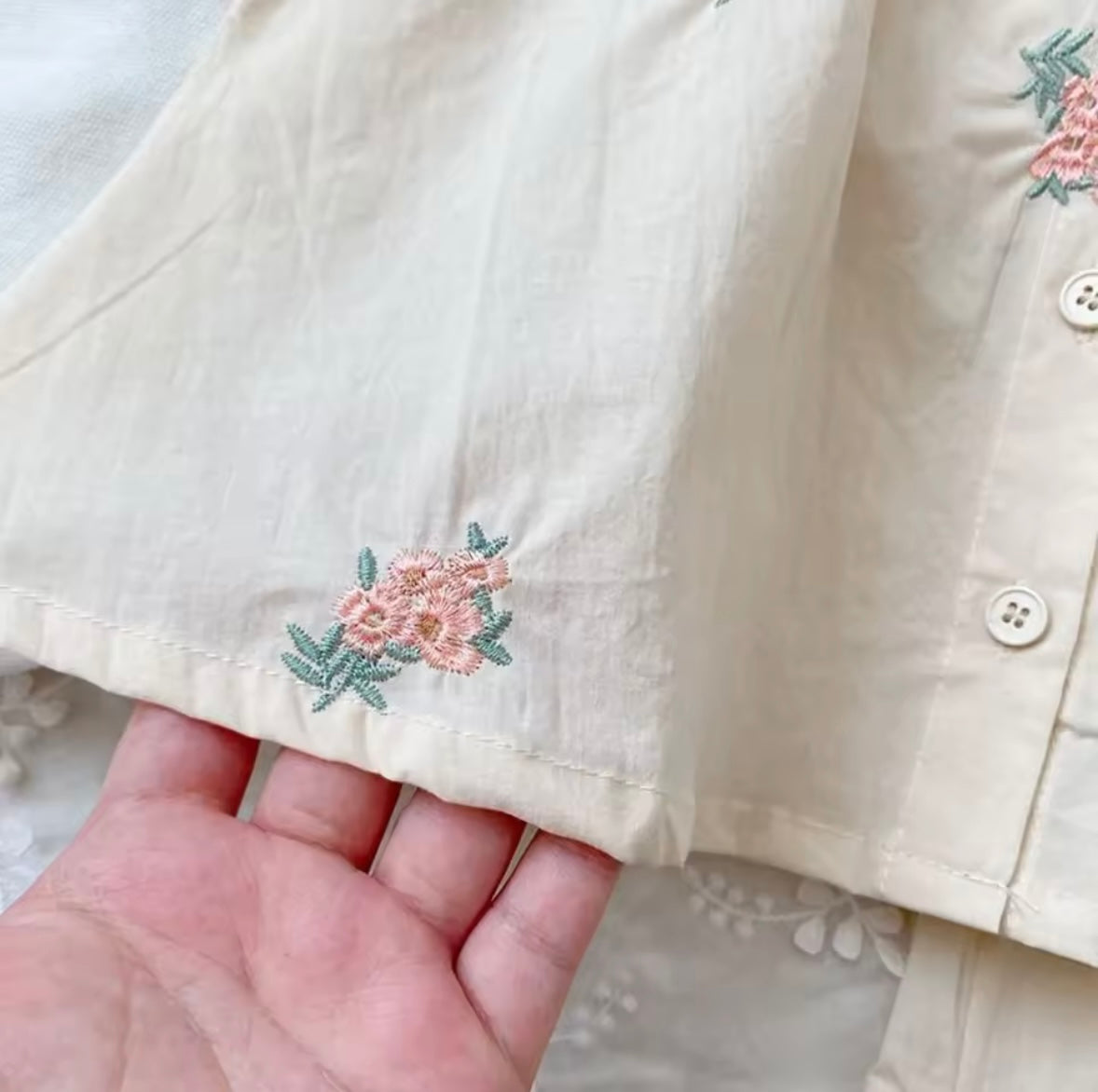 Close-up of a beige shirt with floral embroidery held by a hand.