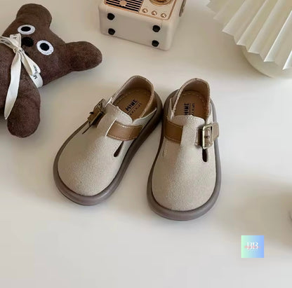 Pair of children's sandals with a teddy bear and toy camera in the background on a white surface
