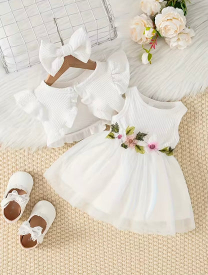 White baby dress with floral details, white shoes, and headband on a textured surface with flowers.