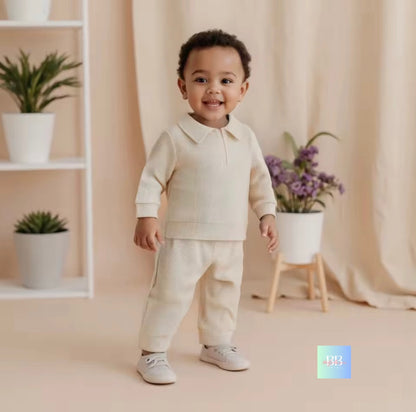 Child wearing a beige outfit standing in a room with plants and a shelf.
