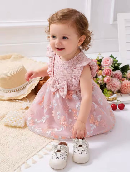 Child in a pink floral dress sitting on a white surface with flowers and a hat in the background.