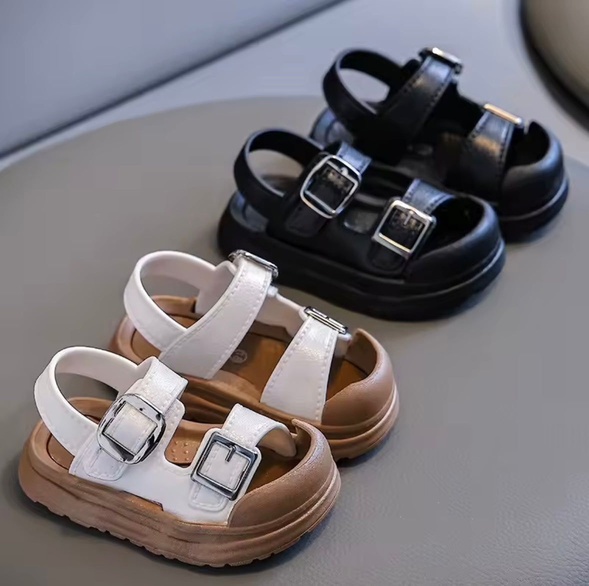 Pair of black sandals and a pair of white sandals with brown soles on a gray surface.