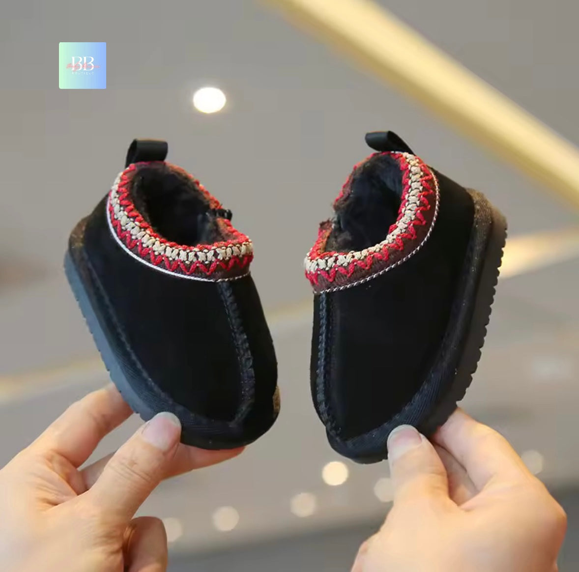 Pair of black children's boots with red and silver trim held by hands against a blurred indoor background.