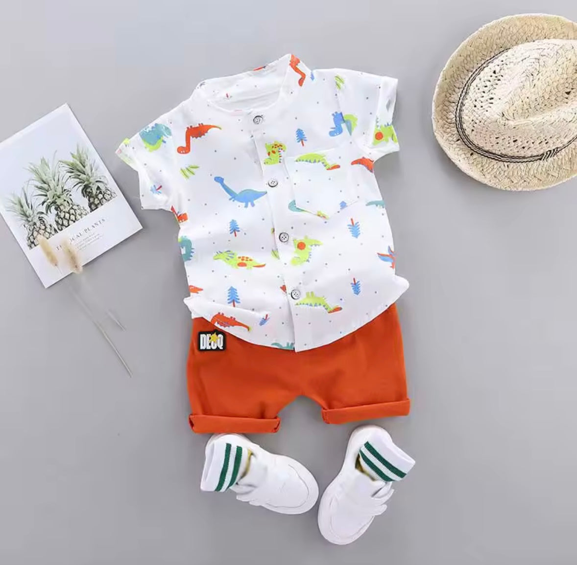 Children's outfit with dinosaur-themed shirt, orange shorts, white socks, and a hat on a gray background.