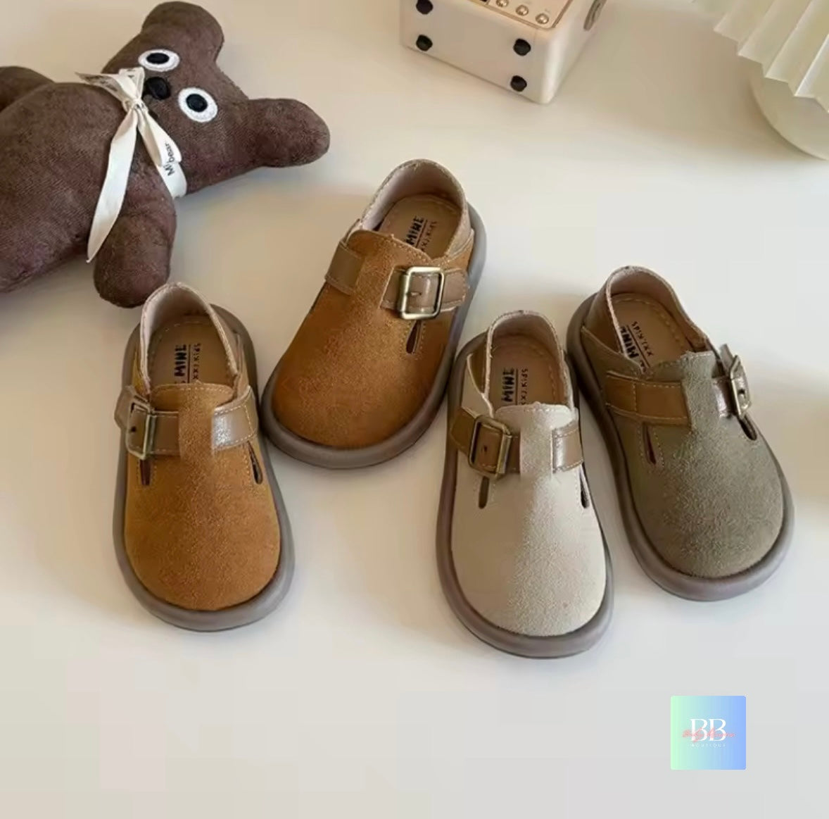 Pair of children's shoes with buckles on a white surface, next to a teddy bear.