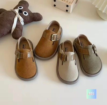Pair of children's shoes with buckles on a white surface, next to a teddy bear.