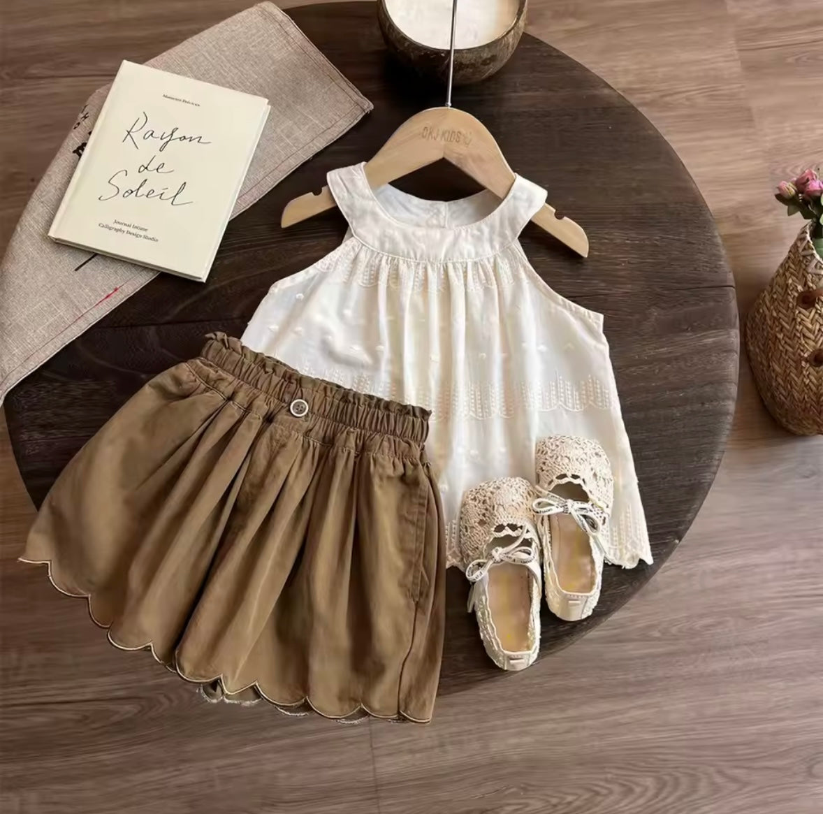 Children's white dress, brown skirt, and shoes on a wooden table with a book and hanger.