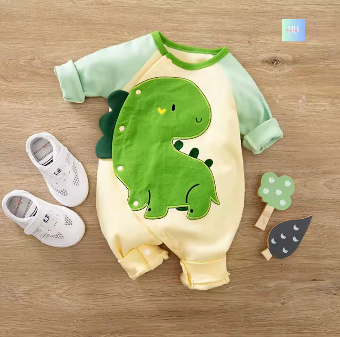 Baby romper with dinosaur design on a wooden surface, accompanied by baby shoes and a toy.