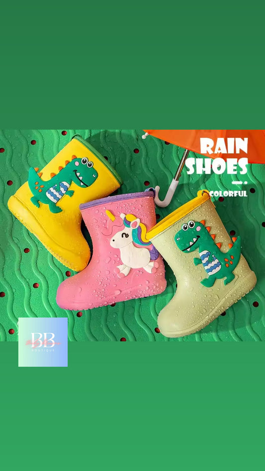 Colorful children's rain boots with dinosaur and unicorn designs on a green textured background.
