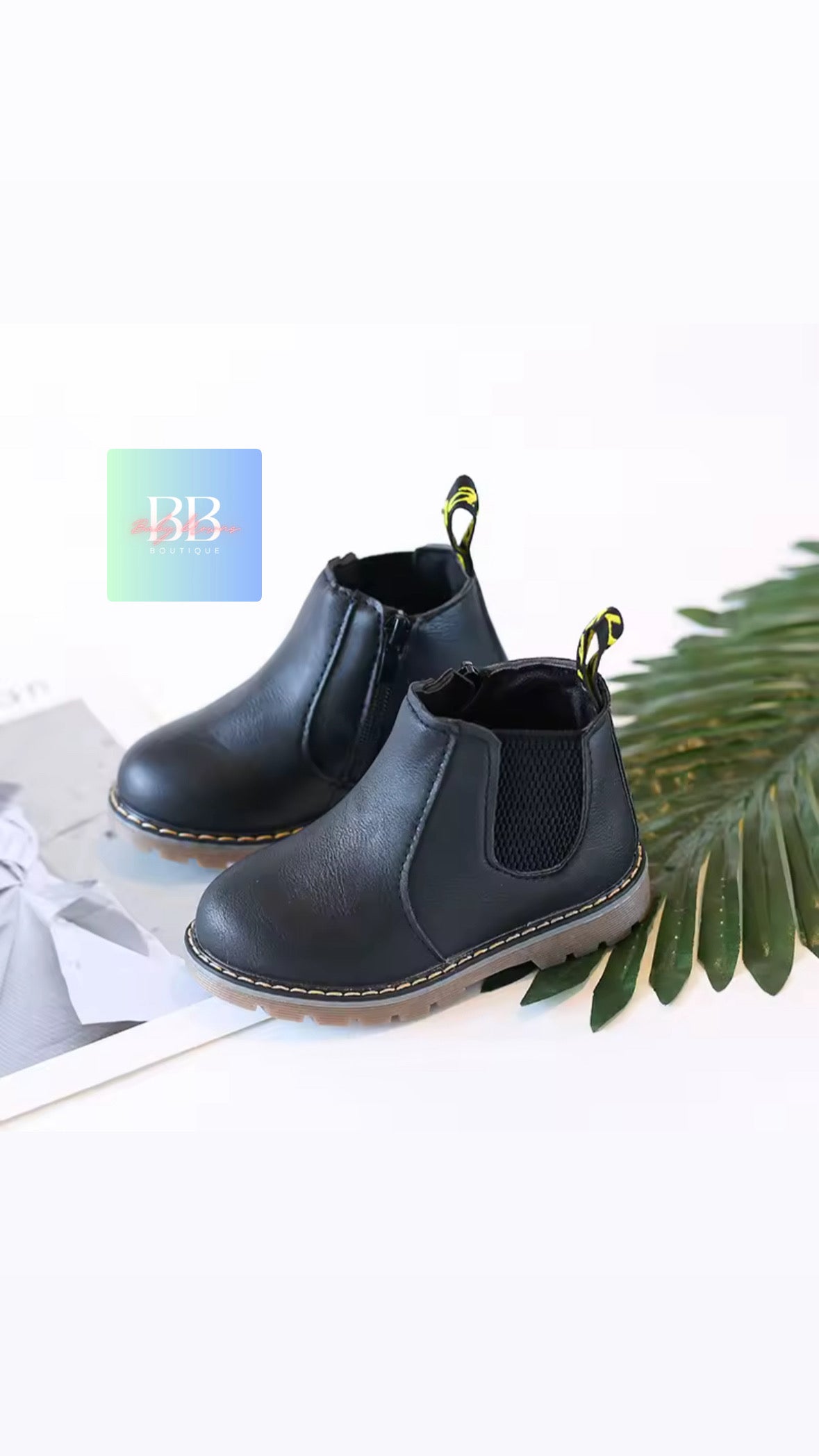 Black Chelsea boots on a white surface with a plant and magazine in the background.