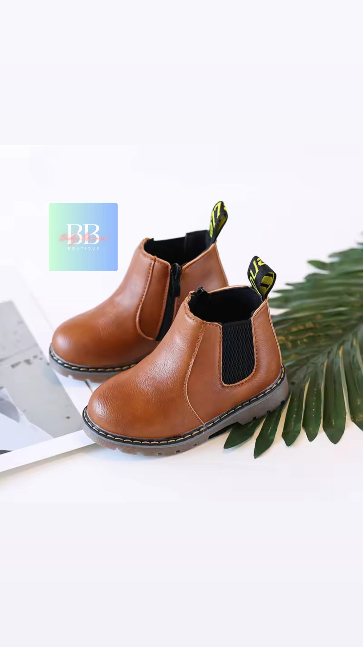 Pair of brown children's boots with black accents on a white surface with a green leaf.