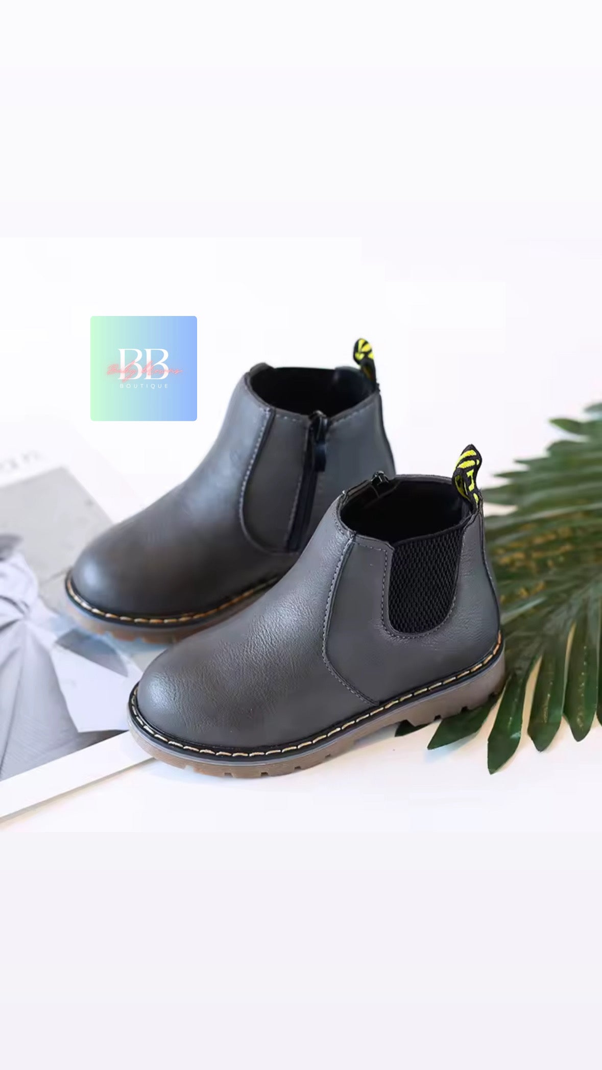 Pair of black Chelsea boots on a white surface with a plant in the background
